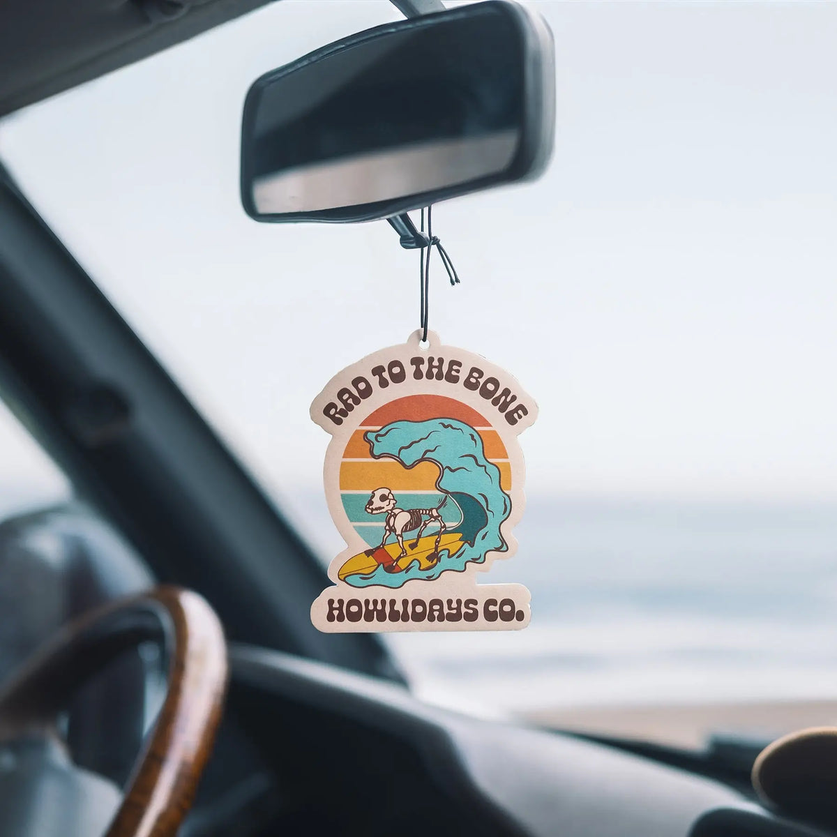 The Rad to the Bone Air Freshener hanging from a vehicle’s rearview mirror with the beach beyond.